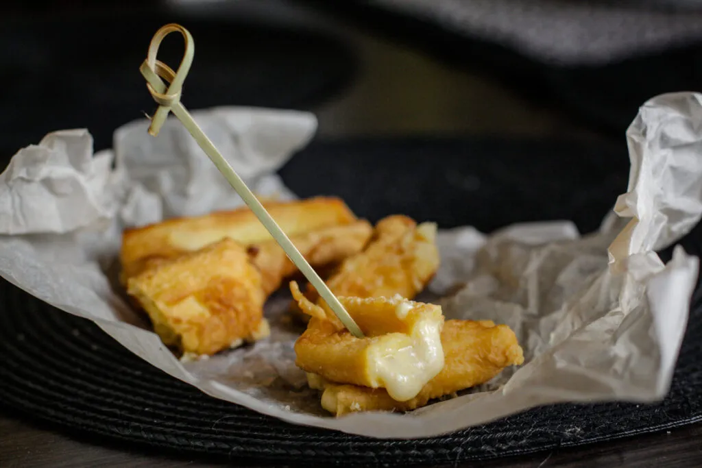 Bocconcini di formaggio fritti in pastella su carta da forno, con il formaggio fuso che fuoriesce da uno dei pezzi infilzato su uno stecchino