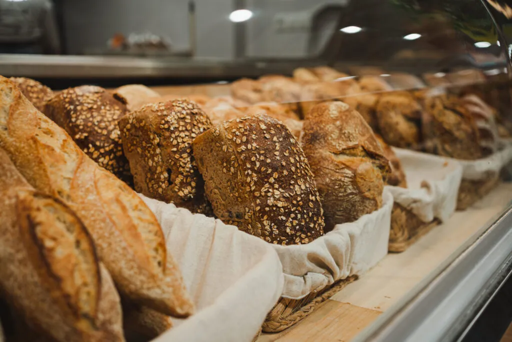 Pagnotte scure ricoperte di semi e fiocchi d'avena accanto a filoni chiari, esposte in un cestino dentro la vetrina di una panetteria