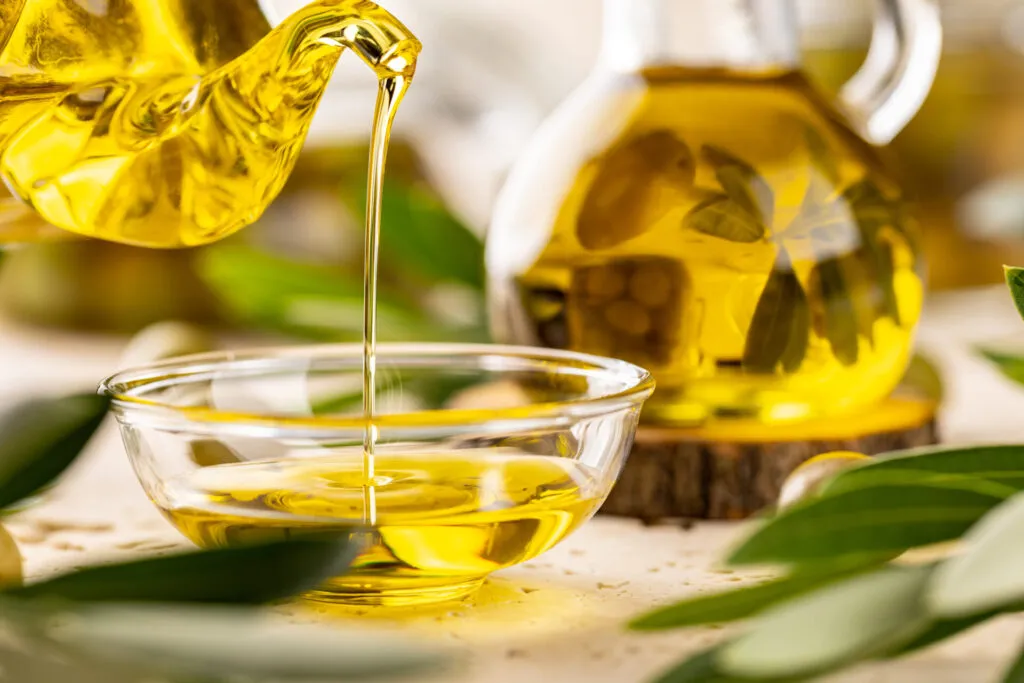 Olio extravergine dal colore dorato versato da un'oliera di vetro in una ciotolina trasparente, con foglie d'olivo intorno