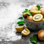 Ciotola di ceramica con kiwi interi e tagliati, due a polpa verde e due spicchi a polpa gialla, con foglie fresche