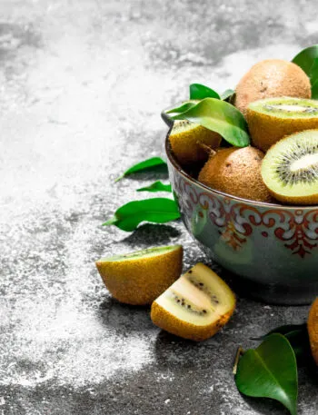 Ciotola di ceramica con kiwi interi e tagliati, due a polpa verde e due spicchi a polpa gialla, con foglie fresche