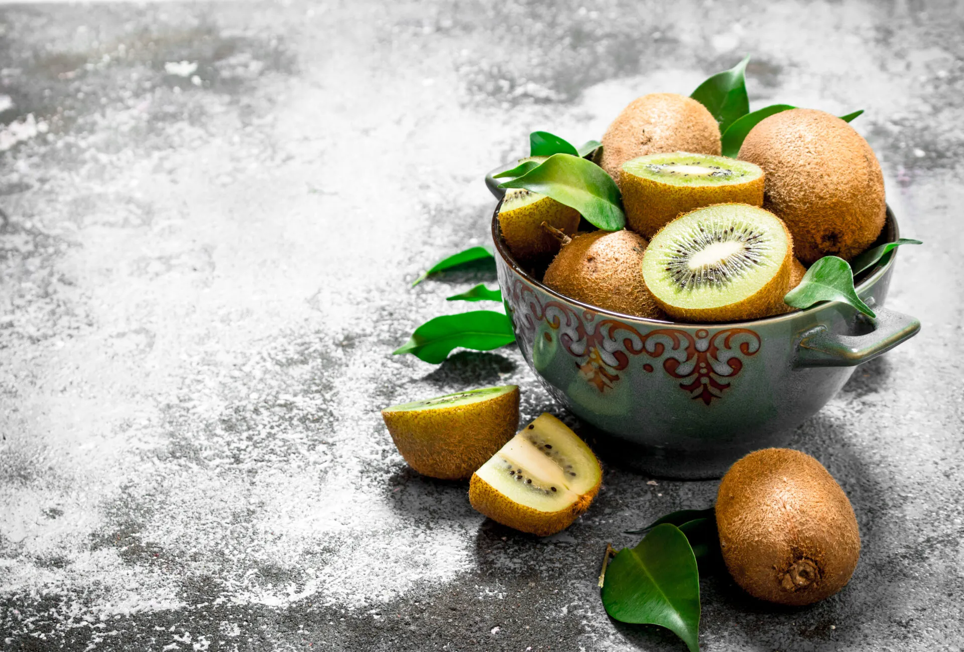 Ciotola di ceramica con kiwi interi e tagliati, due a polpa verde e due spicchi a polpa gialla, con foglie fresche