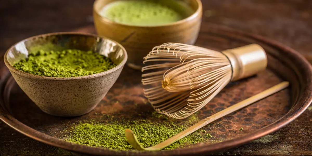 Ciotola di ceramica con polvere di matcha verde, frusta di bambù e cucchiaino su vassoio di rame, con una tazza di matcha preparato sullo sfondo