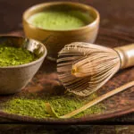 Ciotola di ceramica con polvere di matcha verde, frusta di bambù e cucchiaino su vassoio di rame, con una tazza di matcha preparato sullo sfondo
