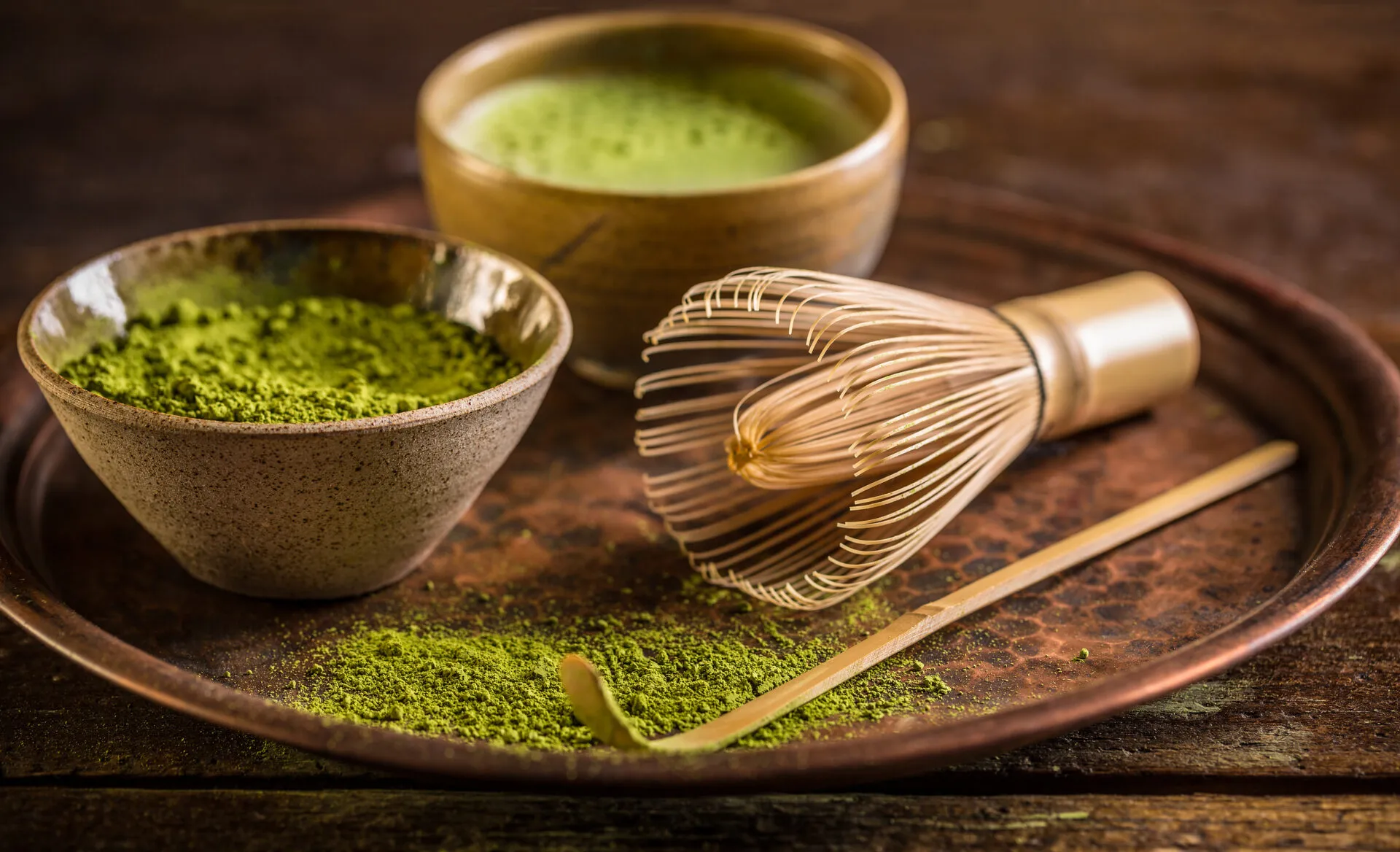 Ciotola di ceramica con polvere di matcha verde, frusta di bambù e cucchiaino su vassoio di rame, con una tazza di matcha preparato sullo sfondo