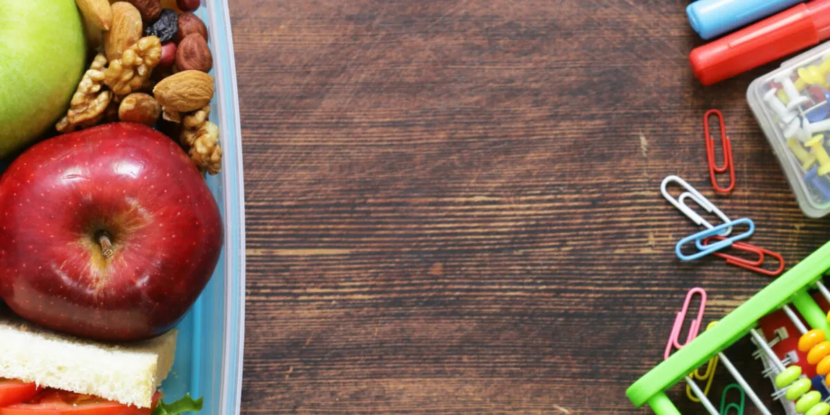 Contenitore per la merenda con mele, frutta secca e un tramezzino, accanto a pennarelli, graffette e un pallottoliere su un tavolo di legno scuro