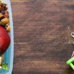 Contenitore per la merenda con mele, frutta secca e un tramezzino, accanto a pennarelli, graffette e un pallottoliere su un tavolo di legno scuro