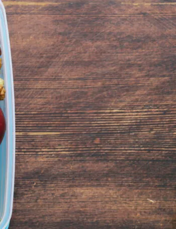 Contenitore per la merenda con mele, frutta secca e un tramezzino, accanto a pennarelli, graffette e un pallottoliere su un tavolo di legno scuro
