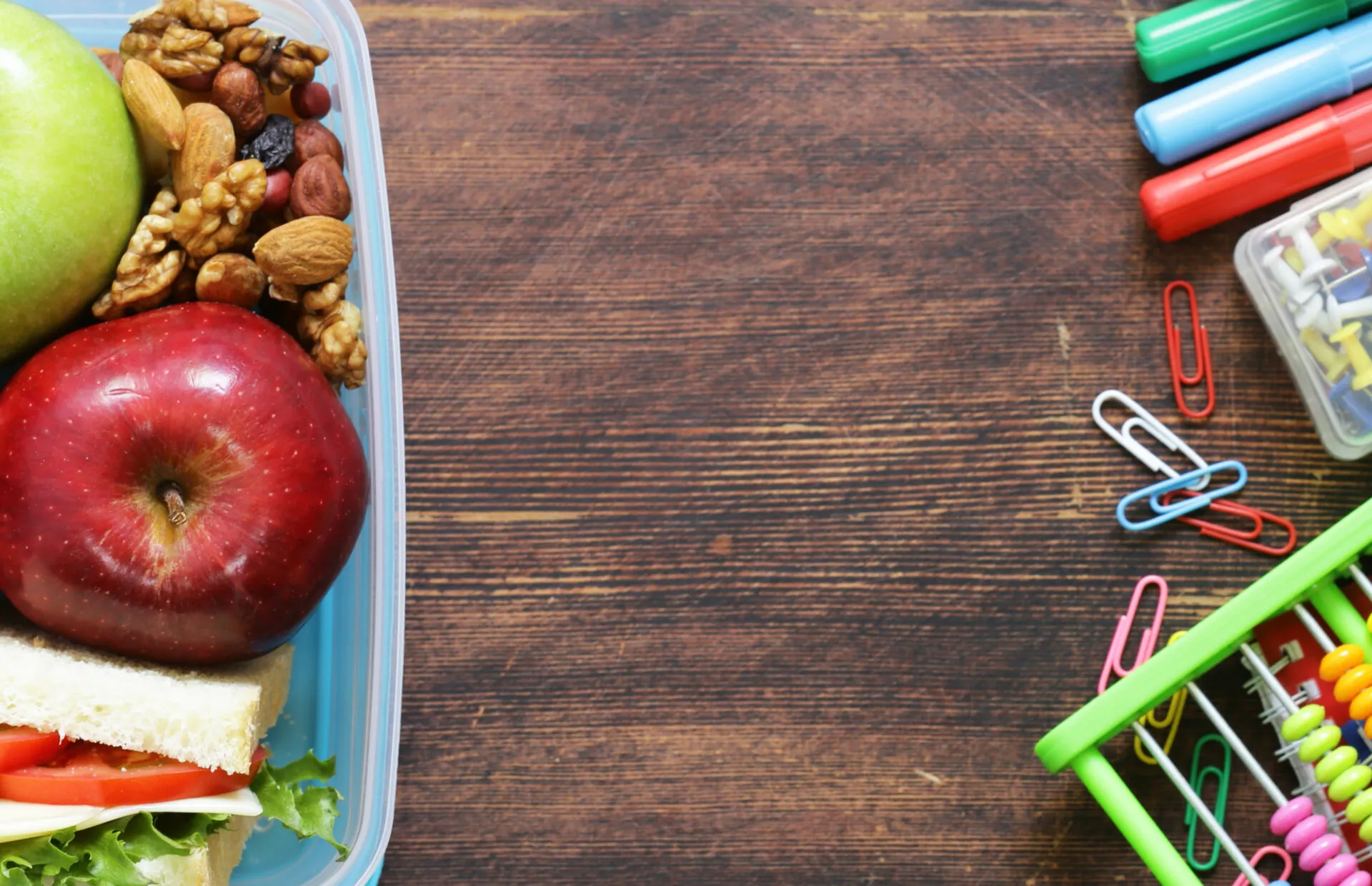 Contenitore per la merenda con mele, frutta secca e un tramezzino, accanto a pennarelli, graffette e un pallottoliere su un tavolo di legno scuro