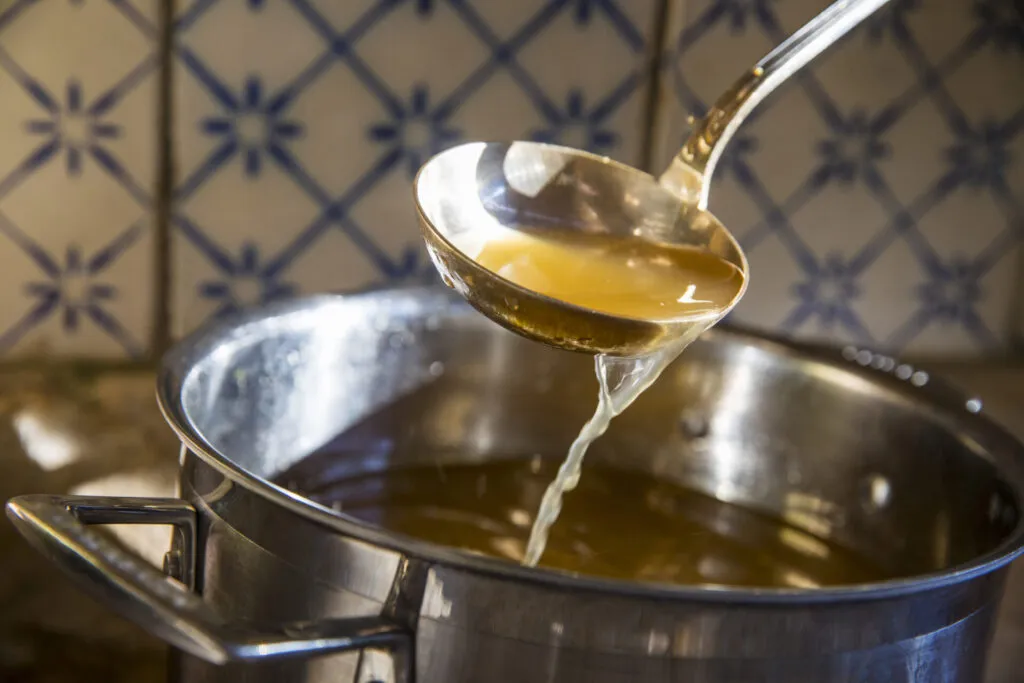Mestolo che solleva brodo vegetale caldo da una pentola di acciaio, con il brodo che ricade a filo