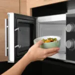 Mano che infila una ciotola verde con verdure cotte dentro un microonde aperto, incassato in un mobile di legno chiaro