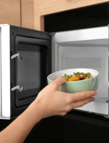 Mano che infila una ciotola verde con verdure cotte dentro un microonde aperto, incassato in un mobile di legno chiaro