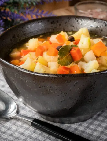 Ciotola nera di minestrone con cubetti di carota e patata, cimette di cavolfiore, una foglia di alloro e grani di pepe, su tovaglia a quadretti grigi con cucchiaio
