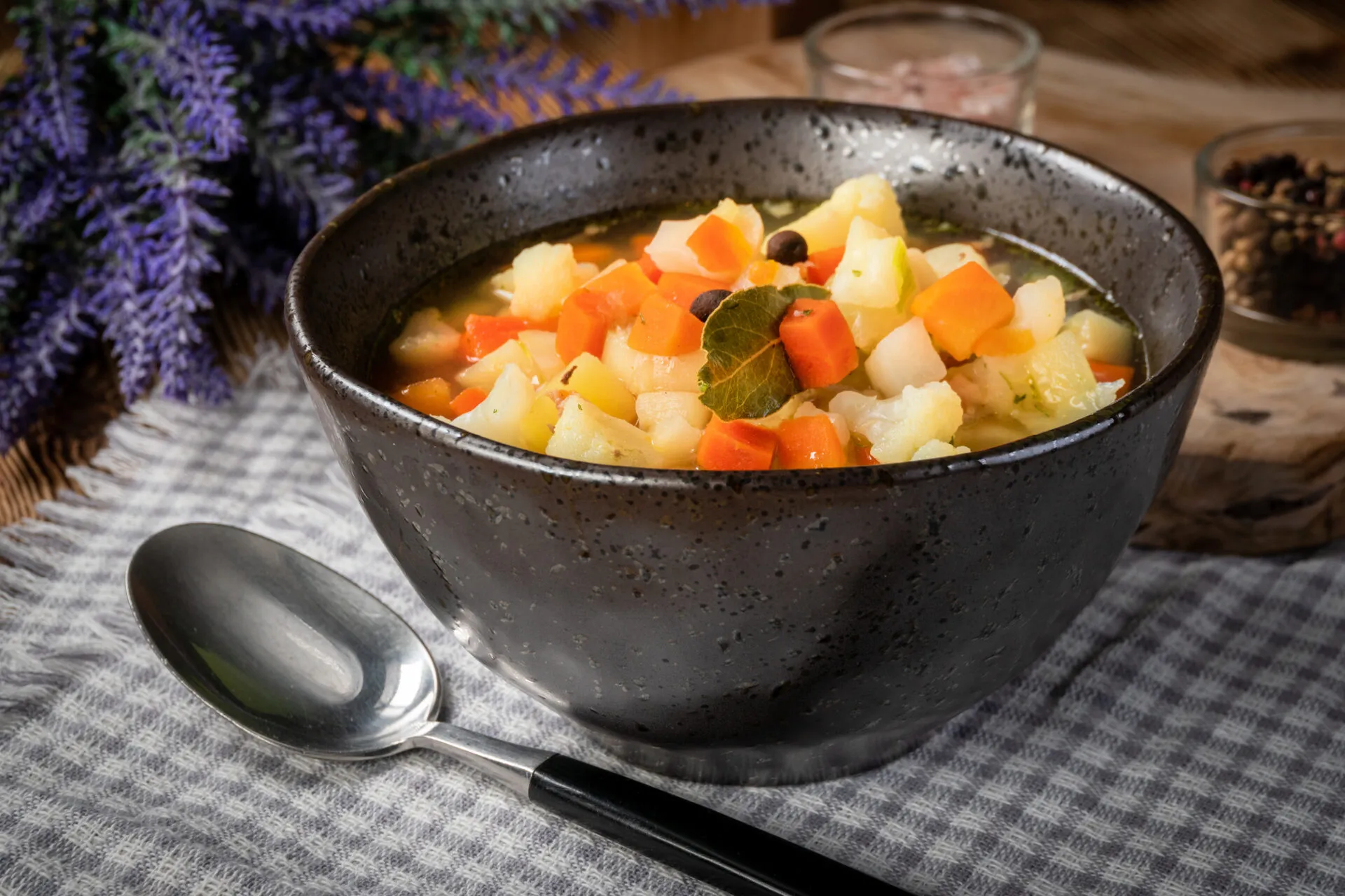 Ciotola nera di minestrone con cubetti di carota e patata, cimette di cavolfiore, una foglia di alloro e grani di pepe, su tovaglia a quadretti grigi con cucchiaio