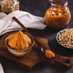 Ciotola di pasta di miso ambrata su un tagliere di legno, con cucchiaio di legno, ciotola di semi di soia e un barattolo sullo sfondo