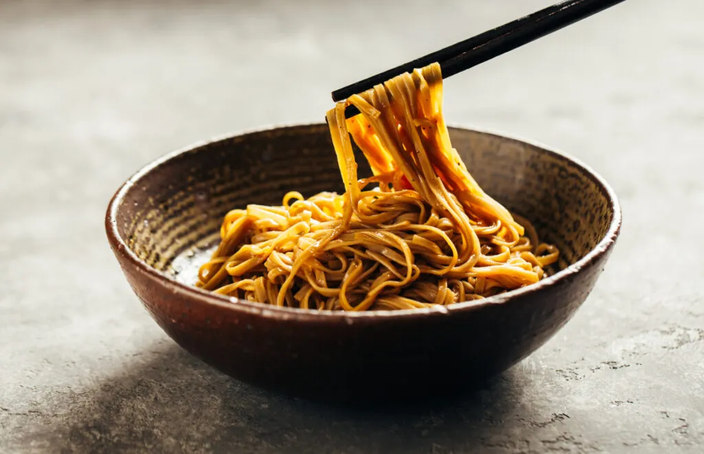 Noodles saltati rivestiti da una salsa lucida sollevati con le bacchette da una ciotola di ceramica