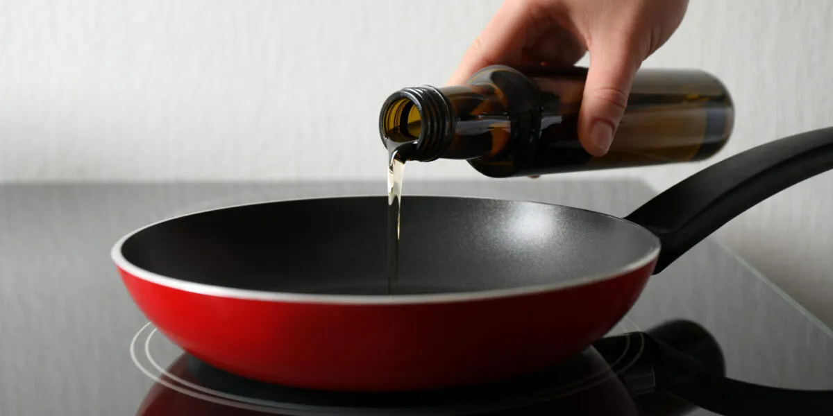 Mano che versa un filo d'olio extravergine da una bottiglia di vetro scuro in una padella rossa antiaderente appoggiata sul piano cottura
