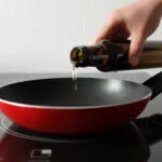 Mano che versa un filo d'olio extravergine da una bottiglia di vetro scuro in una padella rossa antiaderente appoggiata sul piano cottura