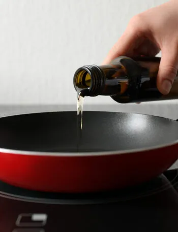Mano che versa un filo d'olio extravergine da una bottiglia di vetro scuro in una padella rossa antiaderente appoggiata sul piano cottura