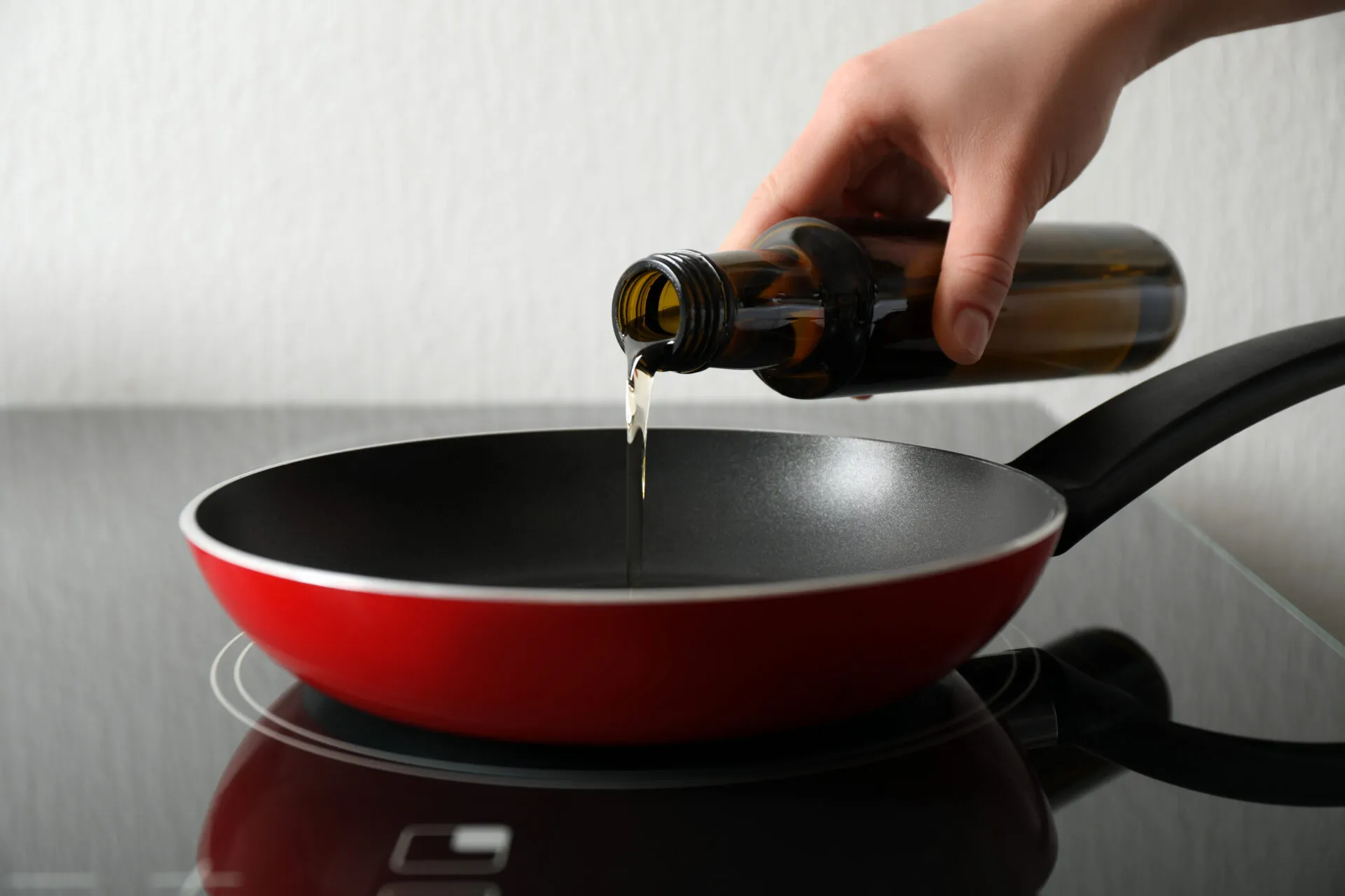 Mano che versa un filo d'olio extravergine da una bottiglia di vetro scuro in una padella rossa antiaderente appoggiata sul piano cottura