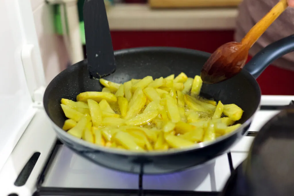 Patate a bastoncino che friggono in padella in uno strato sottile di olio caldo, mescolate con un cucchiaio di legno su fornello a gas