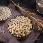 Due ciotole di orzo perlato, una con i chicchi crudi e una con l'orzo cotto e gonfio, con spighe e un barattolo sullo sfondo