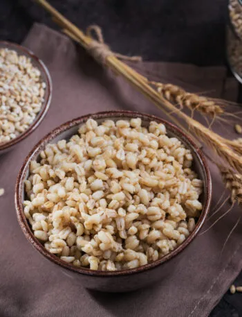 Due ciotole di orzo perlato, una con i chicchi crudi e una con l'orzo cotto e gonfio, con spighe e un barattolo sullo sfondo