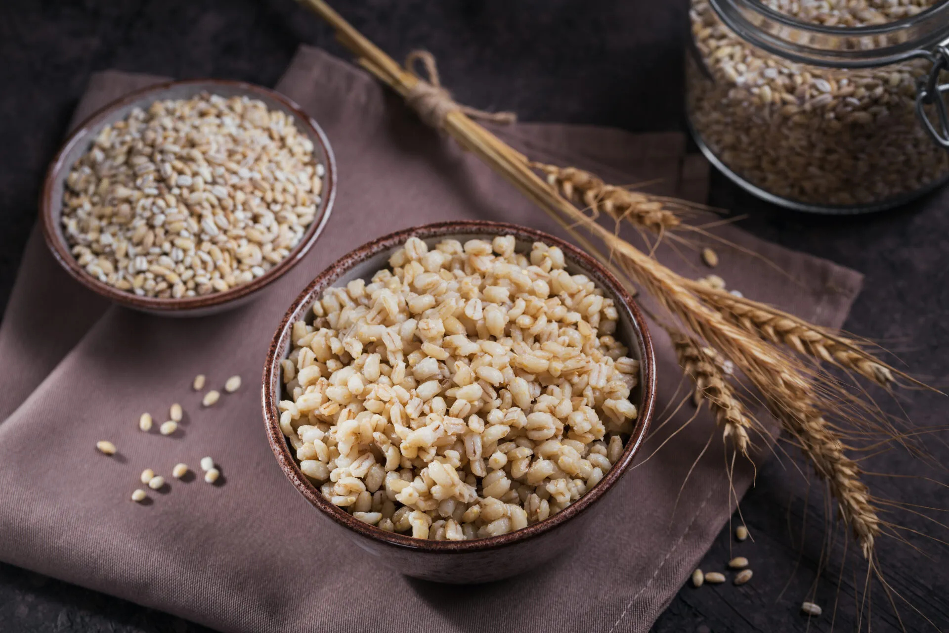 Due ciotole di orzo perlato, una con i chicchi crudi e una con l'orzo cotto e gonfio, con spighe e un barattolo sullo sfondo
