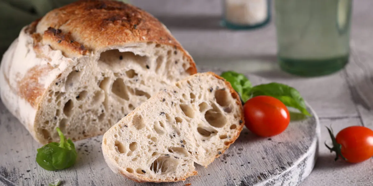 Pagnotta di pane a lievitazione naturale tagliata, con la mollica alveolata e la crosta dorata, su un tagliere di legno con pomodorini e basilico