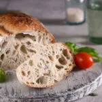 Pagnotta di pane a lievitazione naturale tagliata, con la mollica alveolata e la crosta dorata, su un tagliere di legno con pomodorini e basilico