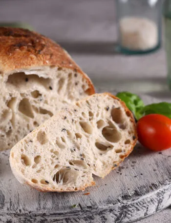 Pagnotta di pane a lievitazione naturale tagliata, con la mollica alveolata e la crosta dorata, su un tagliere di legno con pomodorini e basilico