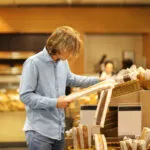 Persona nel reparto pane di un supermercato che osserva una confezione di pane prima di metterla nel carrello, con altri pani confezionati sugli scaffali