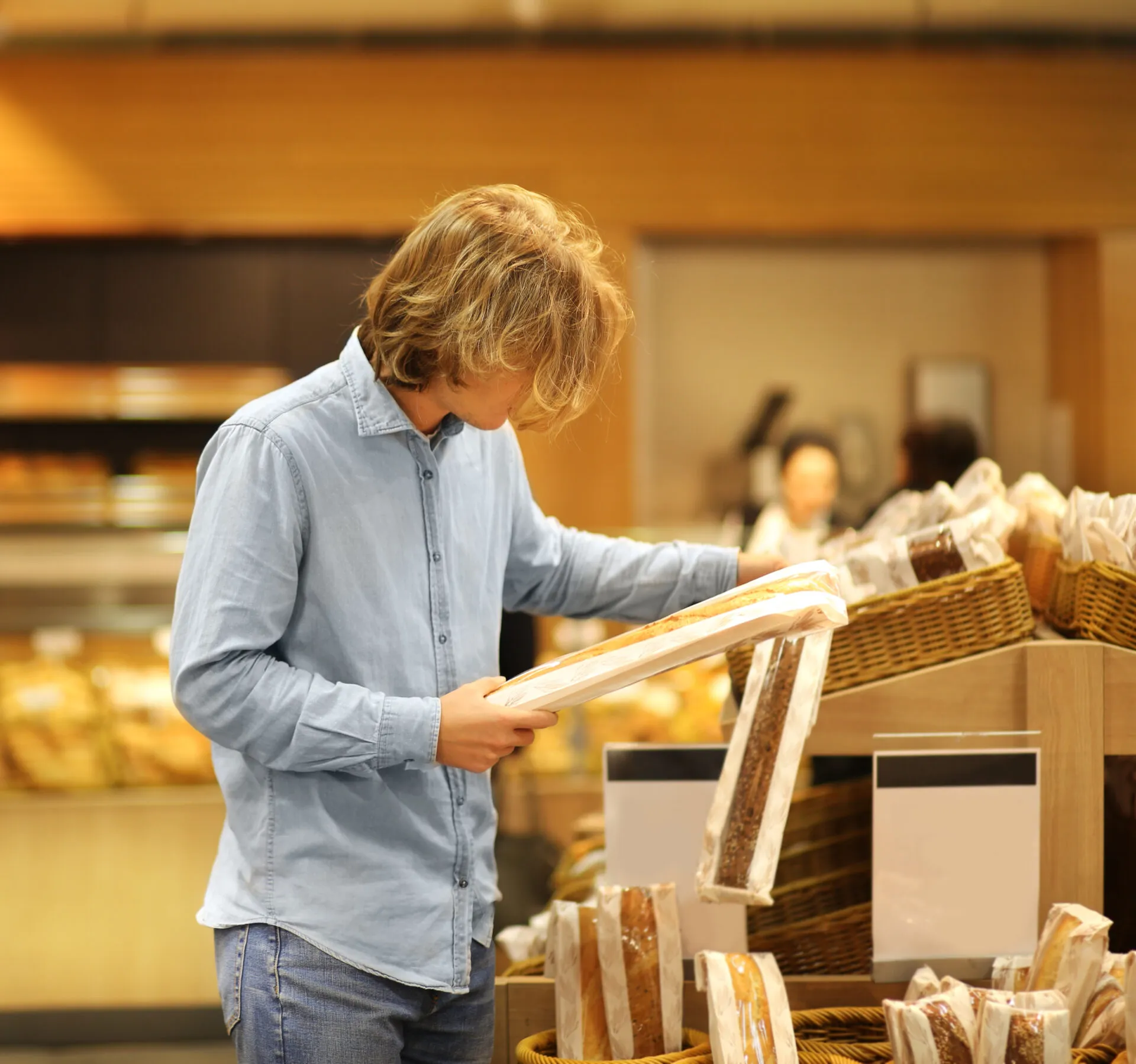 Persona nel reparto pane di un supermercato che osserva una confezione di pane prima di metterla nel carrello, con altri pani confezionati sugli scaffali