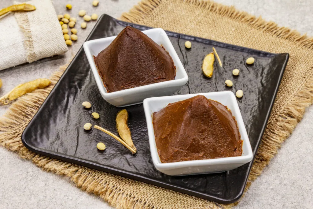 Due ciotoline bianche con pasta di miso scura su un vassoio nero, con semi di soia e baccelli secchi sparsi intorno