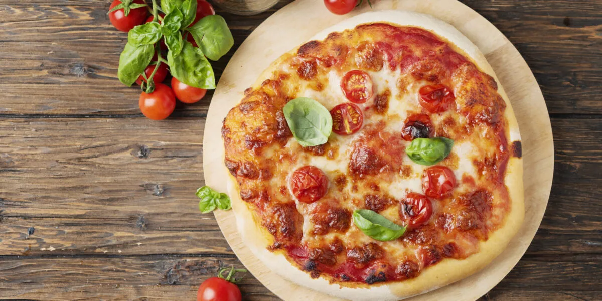 Pizza margherita appena sfornata su un tagliere rotondo di legno, con pomodorini, basilico fresco e un bicchiere accanto