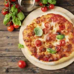 Pizza margherita appena sfornata su un tagliere rotondo di legno, con pomodorini, basilico fresco e un bicchiere accanto