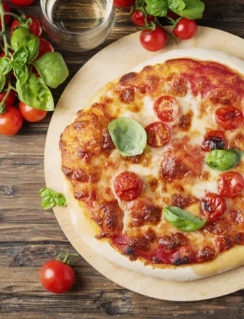 Pizza margherita appena sfornata su un tagliere rotondo di legno, con pomodorini, basilico fresco e un bicchiere accanto