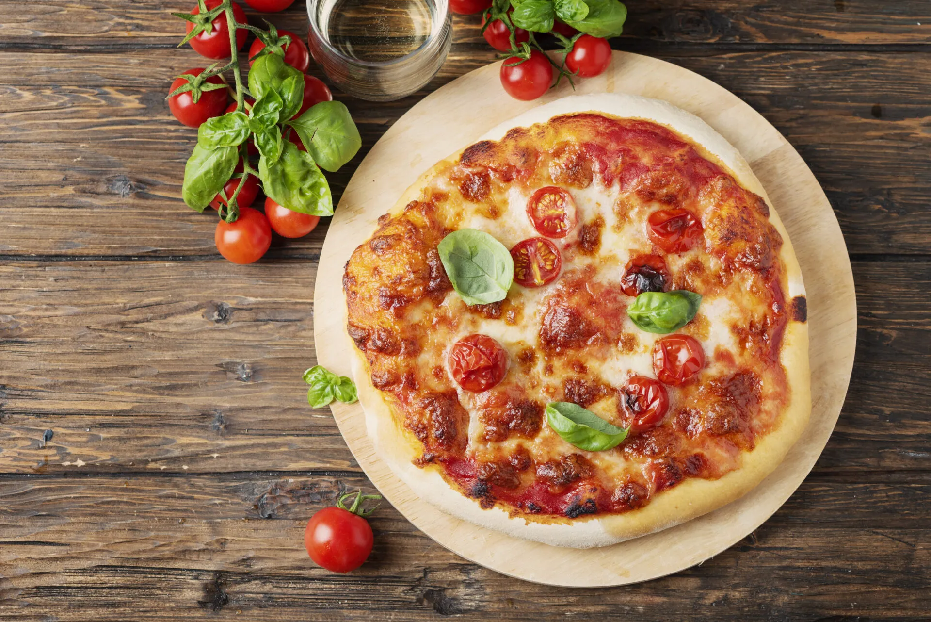 Pizza margherita appena sfornata su un tagliere rotondo di legno, con pomodorini, basilico fresco e un bicchiere accanto