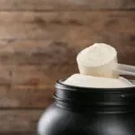 Misurino colmo di proteine in polvere appoggiato sul barattolo aperto, su sfondo di legno