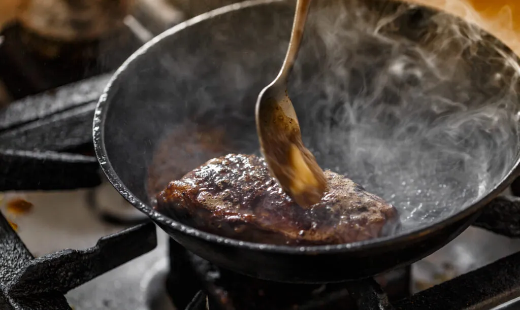 Padella in acciaio su fiamma viva con carne che rosola e fumo denso che sale, mentre un cucchiaio raccoglie il fondo di cottura
