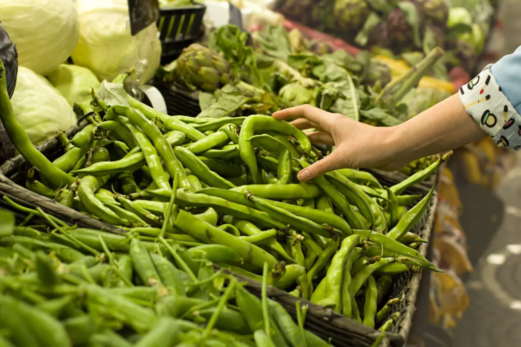Mano che sceglie baccelli di fave fresche da una cassetta al mercato, con carciofi e cavoli sui banchi accanto