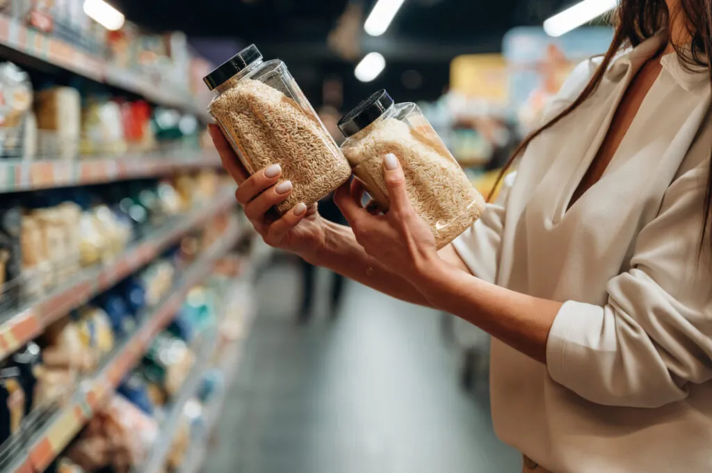 Mani che tengono due barattoli trasparenti di cereali davanti allo scaffale di un supermercato, confrontandoli prima dell'acquisto