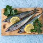 Tre sgombri freschi interi su un tagliere di legno, con le striature scure sul dorso, fette di limone e prezzemolo, su fondo azzurro