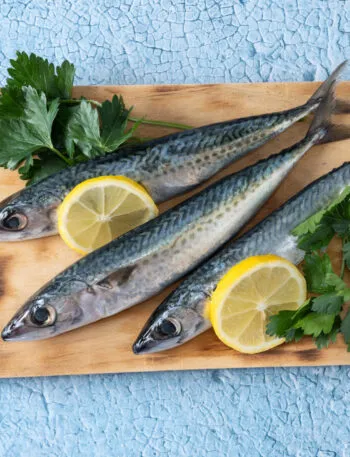 Tre sgombri freschi interi su un tagliere di legno, con le striature scure sul dorso, fette di limone e prezzemolo, su fondo azzurro