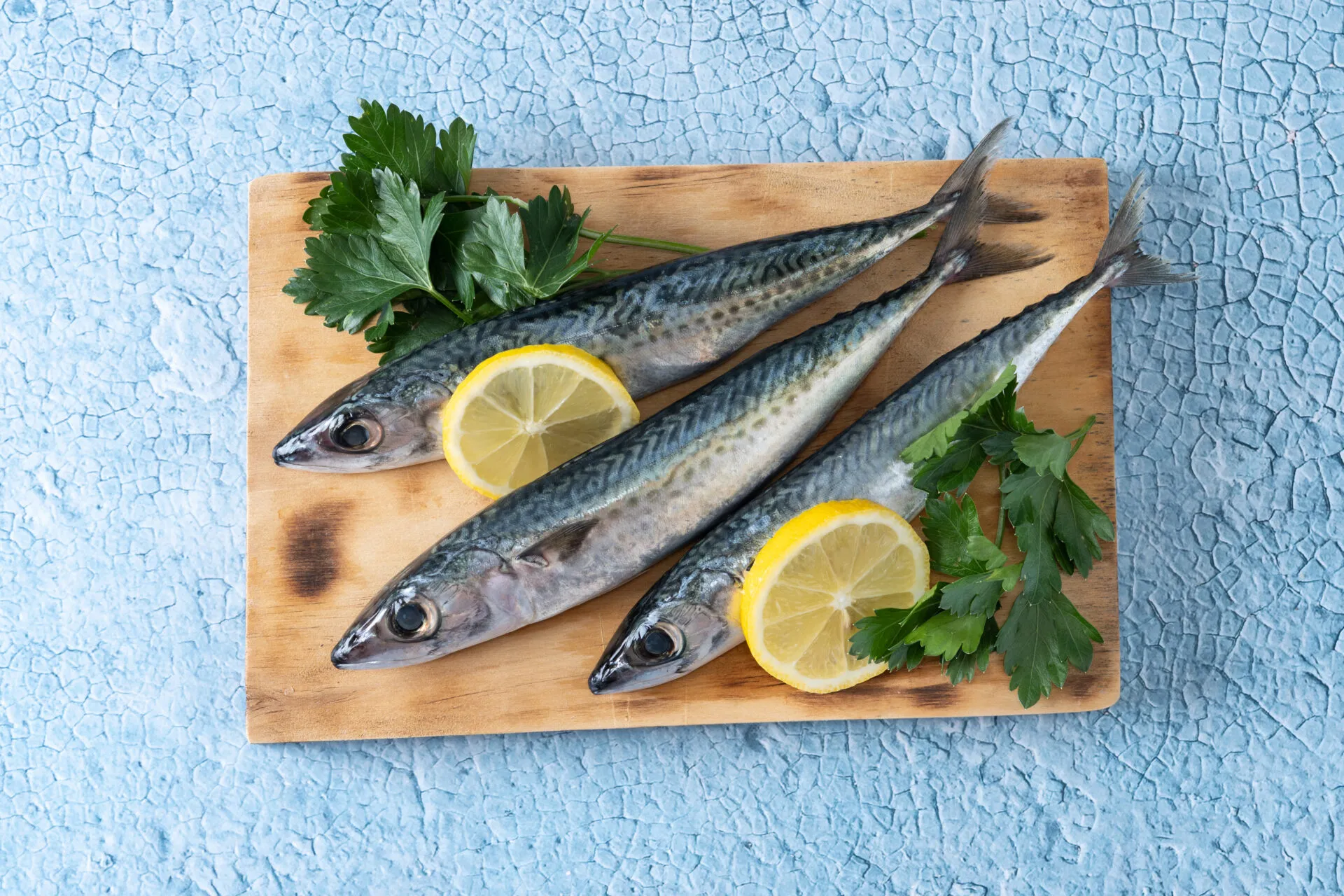 Tre sgombri freschi interi su un tagliere di legno, con le striature scure sul dorso, fette di limone e prezzemolo, su fondo azzurro