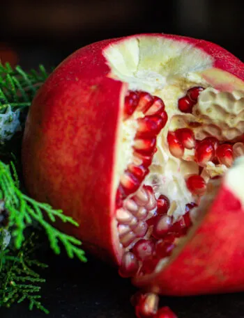 Melagrana rossa aperta a mano lungo le membrane, con arilli rosso rubino e qualche chicco caduto, accanto a rametti di conifera su fondo scuro
