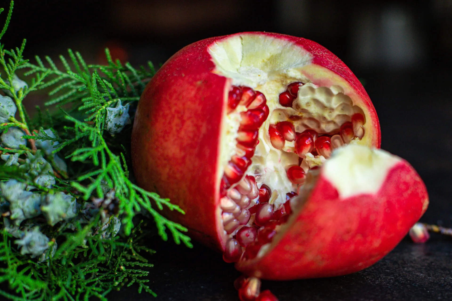 Melagrana rossa aperta a mano lungo le membrane, con arilli rosso rubino e qualche chicco caduto, accanto a rametti di conifera su fondo scuro