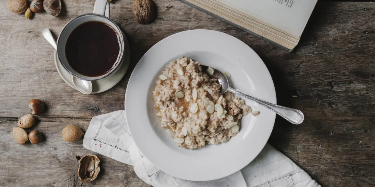 Piatto di porridge d'avena con mandorle a lamelle accanto a una tazza di caffè nero, frutta secca e tovagliolo di lino su un tavolo di legno