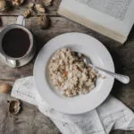 Piatto di porridge d'avena con mandorle a lamelle accanto a una tazza di caffè nero, frutta secca e tovagliolo di lino su un tavolo di legno