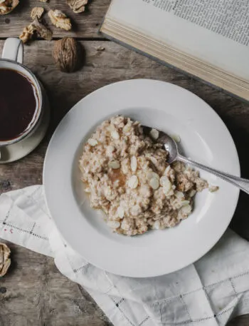 Piatto di porridge d'avena con mandorle a lamelle accanto a una tazza di caffè nero, frutta secca e tovagliolo di lino su un tavolo di legno